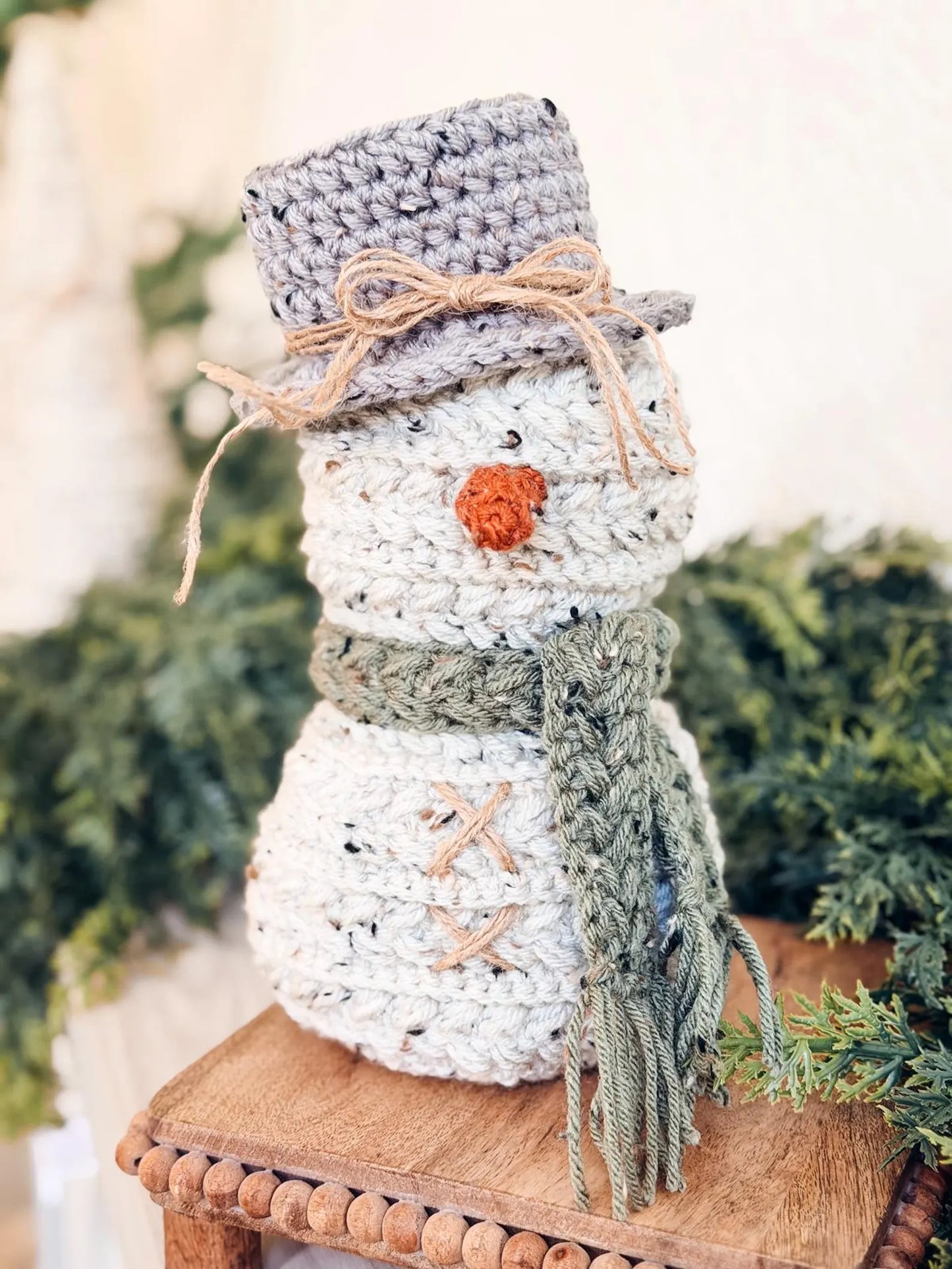 Crocheted snowman decoration with a hat, scarf, and carrot nose on a wooden stool.
