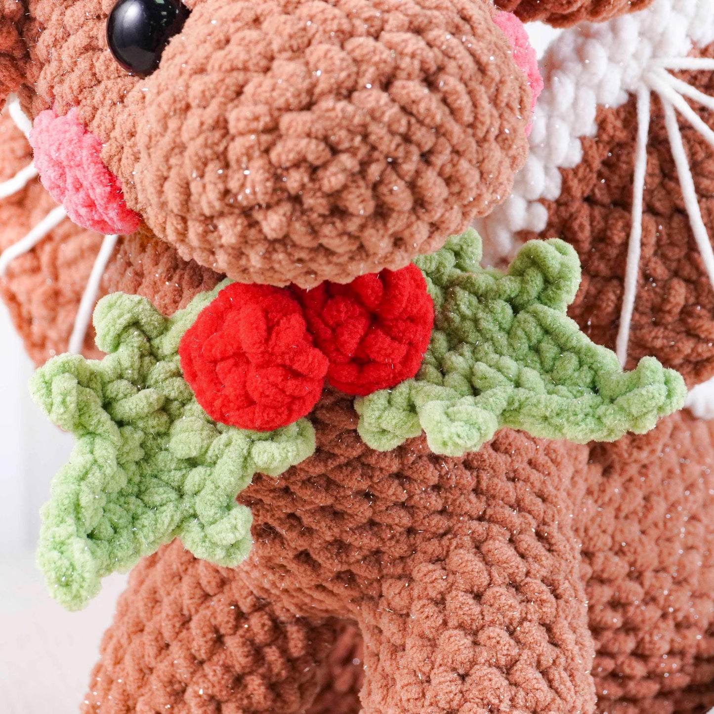 Close-up of a crocheted reindeer with red nose and holly leaves on a white background