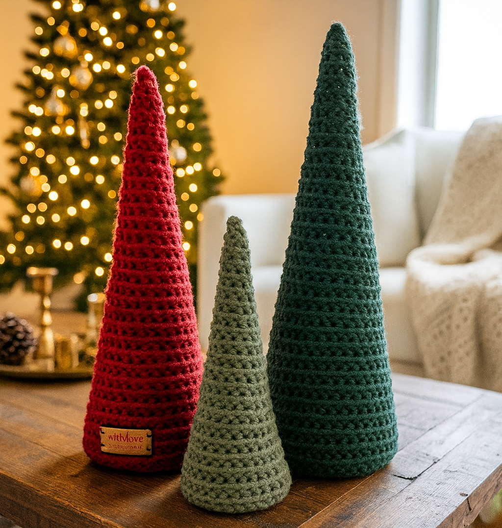 Three crocheted Christmas tree cones in red, green, and brown on a wooden table with a decorated tree in the background.