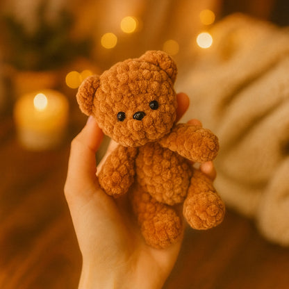 Hand holding a small brown teddy bear against a warm, blurred background with lights.