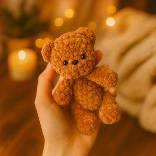 Hand holding a small brown teddy bear against a warm, blurred background with lights.