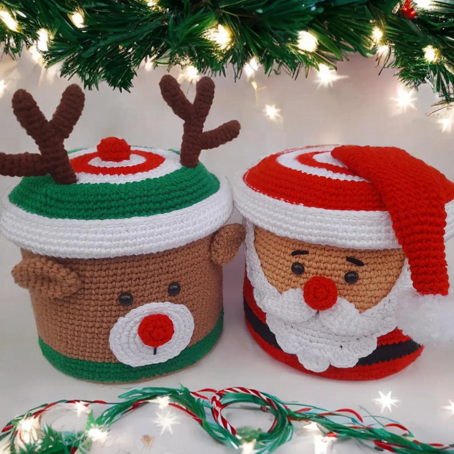 Two crocheted Christmas-themed containers, one resembling a reindeer and the other Santa Claus, with festive lights in the background.