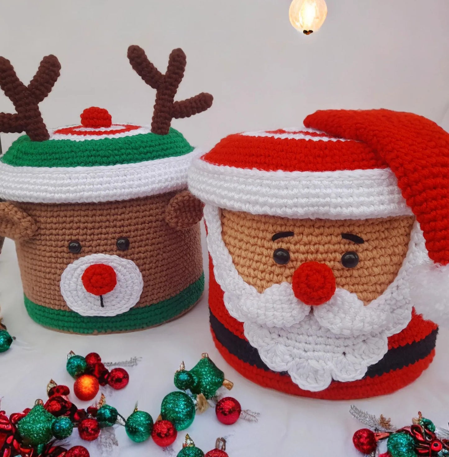 Two crocheted Christmas-themed containers, one resembling Santa Claus and the other a reindeer, with small ornaments on a white background.