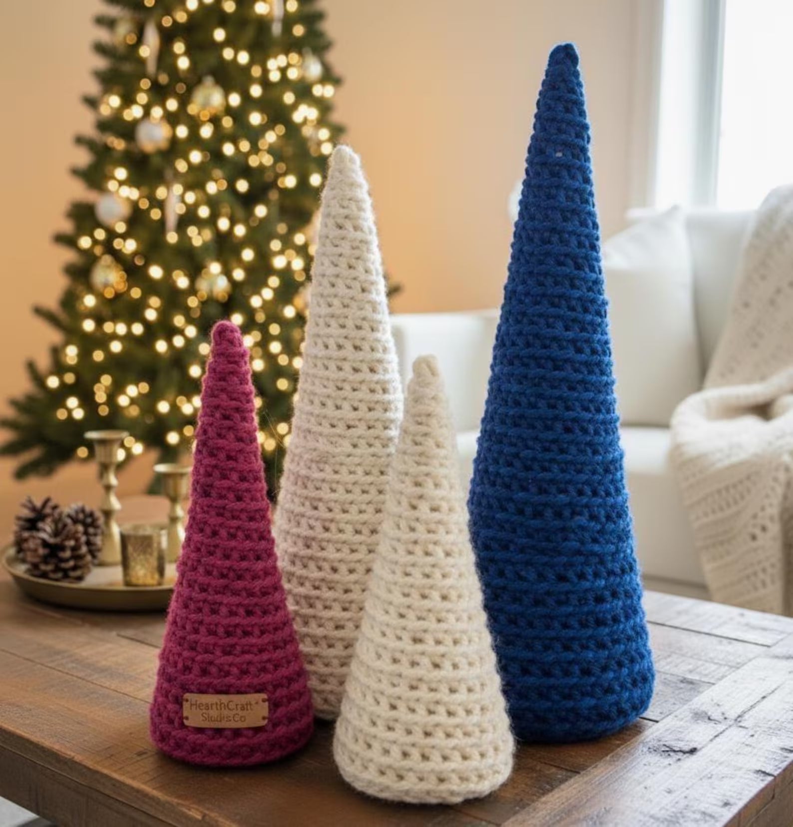 Three crocheted Christmas trees in pink, white, and blue on a table with a decorated tree in the background.