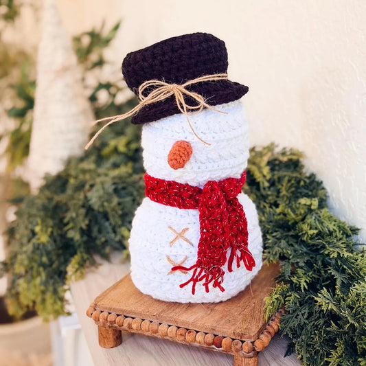 Knitted snowman decoration with a red scarf and black hat on a wooden stand, surrounded by greenery.