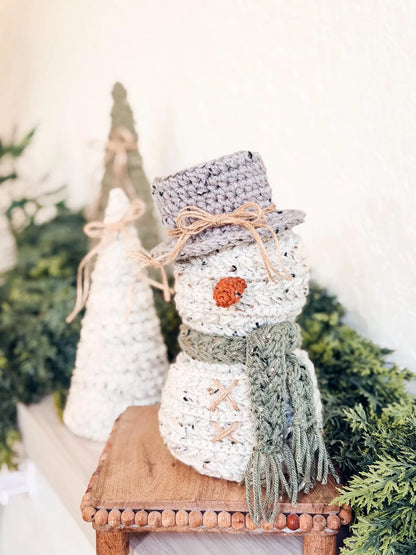 Knitted snowman decoration with a gray hat and green scarf on a wooden stool with Christmas trees in the background.