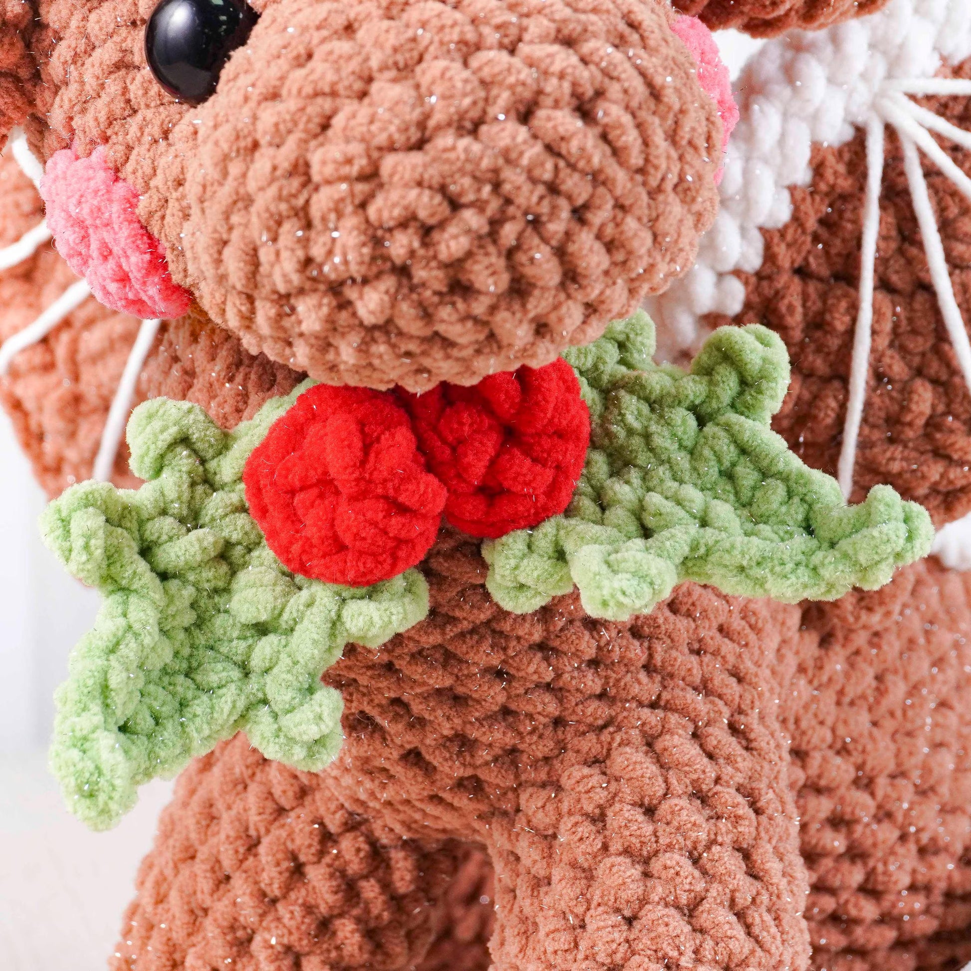Close-up of a crocheted reindeer with red nose and holly leaves on a white background